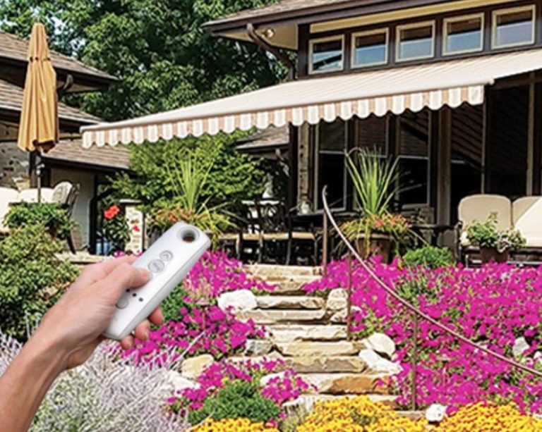 A hand holds a remote control, pointed at a striped retractable awning extended over a patio surrounded by vibrant pink and yellow flowers and greenery.