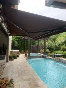 A backyard pool with beige pavers and a large brown motorized awning overhead. Nearby, there are potted plants and a partially covered lounge area. Cascading water features empty into the pool, surrounded by lush greenery and motorized screens for added privacy.