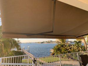 A large, beige retractable awning extends over a patio with a view of a calm waterfront. Palm trees and a manicured lawn are visible near a railing. Buildings are seen across the water under the clear blue sky.