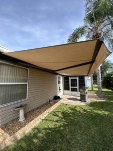A beige retractable awning extends over a patio area next to a house, accompanied by motorized screens for added convenience. Beneath are a grill, potted plant, and decorative pedestal. A well-maintained lawn and tall palm tree stand nearby under the clear, sunny sky.