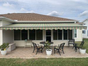 A backyard patio featuring a retractable green and beige striped awning with motorized screens. Beneath it, find a black metal table with chairs, a bench, and a small side table on the brick surface. White planters with plants are tastefully arranged around the space.