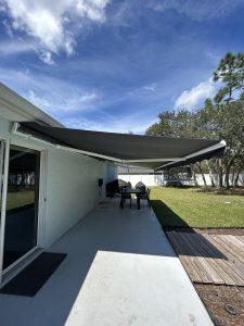 A patio with a retractable awning extends from the house, complemented by motorized screens. A table with chairs sits on the concrete surface, bordered by grass. Trees and a clear blue sky frame the tranquil setting perfectly.