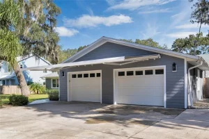 A modern two-car garage with blue siding and white trim features two white overhead doors with small windows at the top. The driveway is wide and paved, complemented by motorized awnings. Surrounding the garage are trees and greenery, beneath a bright, clear sky.