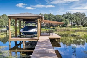 A boat is docked under a wooden shelter on a calm lake. The dock stretches over the water with a patio area featuring a table and chairs beneath motorized awnings. Trees encircle the lake under a blue sky with scattered clouds.