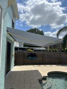 A backyard with a gray motorized retractable awning over a patio beside a small pool. The sky is partly cloudy, and a wooden fence lines the perimeter. Houses and a tree are visible in the background.