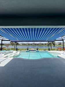 A rectangular swimming pool with steps and clear water beneath motorized awnings that offer a wide blue-and-white striped cover. Beyond the pool, you enjoy a scenic view of the lake, lush greenery, and distant buildings in a bright and sunny atmosphere.