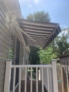 A striped motorized awning gracefully extends over a small outdoor balcony attached to a beige house. The balcony features white railing, and trees are visible in the background under a clear blue sky.