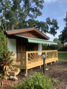 Small wooden cabin with a green awning, partially covered deck, and decorative railing. Surrounded by lush greenery and trees under a clear blue sky. A garden lamp stands in front on mulch-covered ground, alongside various plants and retractable awnings for effortless shade.