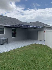 A backyard patio features a striped blue and white motorized retractable awning attached to a gray house with two windows. The speckled gray surface is bordered by a lush green lawn, complete with an air conditioning unit and a white fence in view.