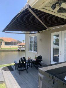 Two black Adirondack chairs with small tables sit on a deck under a motorized retractable awning. The deck overlooks a calm canal with houses across the water. The area is bathed in sunlight, and the sky is clear and blue.