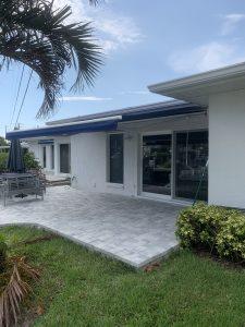 A backyard view of a single-story house features sleek white walls and sliding glass doors. A blue motorized awning shades the patio, which has light gray paving stones. The outdoor furniture sits beside a closed umbrella amid lush green grass and plants in the foreground.