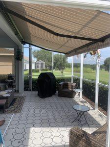 A patio with hexagonal tile flooring showcases wicker furniture, a covered grill, and a retractable striped awning. The area is enclosed with motorized screens, offering a scenic view of greenery and trees in the background.