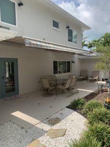 A patio features a long outdoor dining table and chairs under a retractable awning attached to a two-story house. The space is adorned with potted plants, green shrubs, and a stone pathway, all set against the backdrop of a cloudy sky.