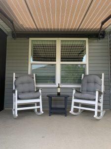 Two white rocking chairs with gray cushions face each other on a porch. A small black table between them holds a wine bottle. The retractable awning above provides shade, while reflections in the window reveal glimpses of the roof and sky.