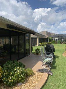 The patio area, featuring motorized awnings over reclining chairs and lush plants, is adjacent to a grassy lawn. A barbecue grill stands covered nearby. Behind, houses boast screened-in porches under a partly cloudy sky.