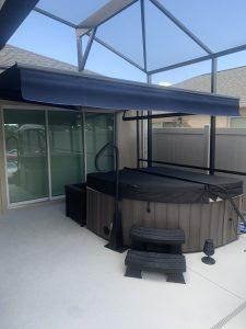 A covered hot tub graces the white patio floor, nestled under motorized retractable awnings and surrounded by sleek glass walls. Steps lead up to the inviting water, with a handy storage box nearby for added convenience.