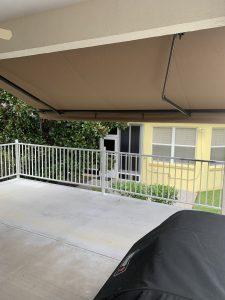 A patio area with a motorized retractable awning offers shade. The space features white railings and overlooks a garden. In the foreground, theres a covered barbecue grill. In the background, a yellow building with windows and a door is visible.