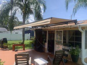 A backyard patio features a beige motorized awning extending over the seating area, with chairs, a white table, and potted plants. A palm tree and green lawn are visible, framed by a white fence under a clear blue sky.