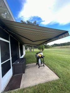 A striped retractable awning extends over a concrete patio with green grass surrounding it. Underneath, theres a grill and a storage box. The sky is blue with some clouds, and trees are visible in the distance.