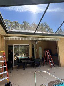 Patio area with a glass roof, two red ladders, and outdoor furniture. A ceiling fan is visible above the table. A pool is in the foreground, with a person in dark clothing standing in the background near a glass door.