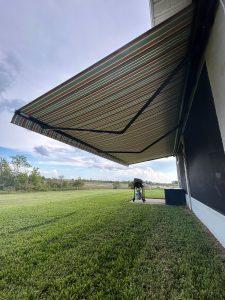A wide, striped awning extends over a patio, providing shade. Behind the awning is a barbecue grill and a small storage box. The view includes a grassy area with trees and a partly cloudy sky in the background.
