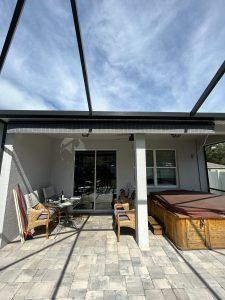 Patio with wicker chairs and a set table on the left, a closed hot tub on the right, and a glass door in the center. The area is covered by a metal frame overhead. The sky is partly cloudy.