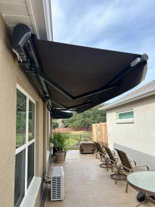 Patio area with a retractable awning attached to a house. Beneath the awning, there are outdoor chairs, a table, potted plants, and a small air conditioning unit. The sky is partly cloudy.