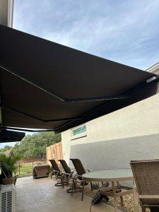 Outdoor patio with a large, dark retractable awning providing shade. Beneath it are several chairs around a round table. A potted plant and grill are nearby, with a view of a fenced yard under a partly cloudy sky.