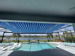 View of a patio with a swimming pool under a blue and white striped awning. The area is furnished with chairs and overlooks a grassy yard with a pond and houses in the background. The sky is partly cloudy.