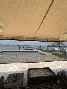 View from under a patio awning overlooking a pool and waterfront dock. The scene includes wicker patio furniture, a tiled deck, and a palm tree, with a calm blue sky and rippling water in the background.