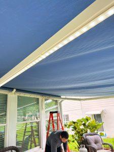 A person is setting up a patio area with a blue fabric awning overhead, equipped with LED strip lights. A red ladder is nearby, and theres greenery and patio furniture in the background.