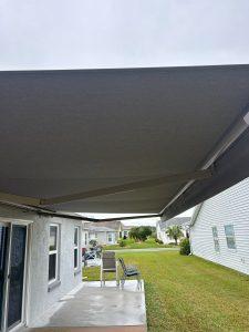 A large gray patio awning extends over a concrete patio beside a white house. Four gray patio chairs are arranged around a glass table. The sky is overcast, and a green lawn extends to another white house in the background.