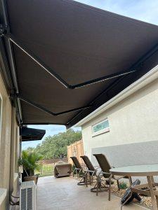 A covered patio with reclining chairs and a table underneath a large dark retractable awning. The patio features a light-colored wall with a small window and overlooks a yard with trees and a fence.