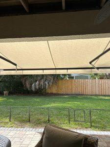 A backyard view with a cream-colored awning extending over a patio area. Theres a lawn enclosed by a wooden fence, with trees visible in the background. Wicker chairs with cushions are situated in the foreground.