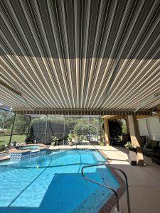A covered swimming pool area with striped awnings casting shadows on the water. Theres a seating area to the right and potted plants around the pool. The space is enclosed with glass screens, showing a view of greenery outside.