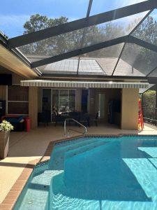 A screened outdoor area features a swimming pool with clear water. Theres a shaded patio with chairs and a few items in the background. The structure has a sloped roof with solar panels on top, surrounded by trees and a clear blue sky.