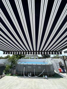 A backyard scene with a large, black and white striped canopy overhead, providing shade to a round, above-ground pool with metal supports. The pool area is surrounded by a white fence, with various plants and outdoor accessories nearby.