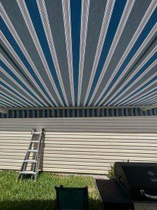A view from beneath a blue and white striped awning, looking out at a sunlit wall with a metal ladder leaning against it. A black grill is partially visible on the right, and green grass is at the bottom.
