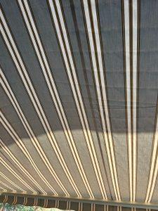 View of a fabric awning with vertical stripes in shades of brown, cream, and green. Sunlight casts shadows on the material, highlighting the pattern. Part of the sky is visible at the bottom edge.