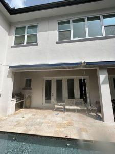 A modern white two-story house with a backyard patio. Features include a grill, swing bench, and a small pool in the foreground. Large windows and doors provide natural light, while an awning offers shade over the seating area.