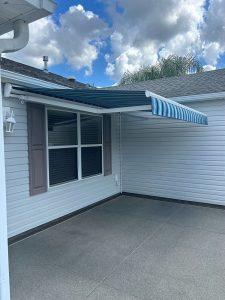 A blue and white striped retractable awning extends over a patio area attached to a house. The building exterior is light-colored with a window and shutters. The sky is partly cloudy, and a palm tree can be seen in the background.