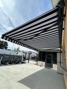 A patio with a striped black and white retractable awning attached to a building. The area features a swimming pool, a white fence, and clear blue sky above. A black door and windows are visible on the house wall.
