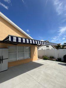 Side view of a beige house with a black and white striped awning extending over a large window. The awning casts a shadow on the concrete patio. A white fence and some greenery are visible in the background under a clear blue sky.
