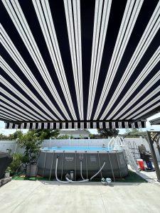 A backyard with an above-ground swimming pool under a black-and-white striped canopy. The pool is surrounded by a privacy fence and some greenery. The ground is paved, and a ladder leads into the pool.
