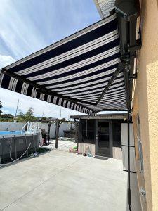 A striped retractable awning extends over a backyard patio, providing shade near a beige house. The area includes a pool and outdoor furniture. The sky is clear and sunny.