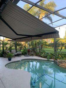 A backyard pool area with a curvy swimming pool, surrounded by lush plants and palm trees. Theres a striped retractable awning providing shade and outdoor seating including chairs and a table. The area is enclosed by a screened structure.