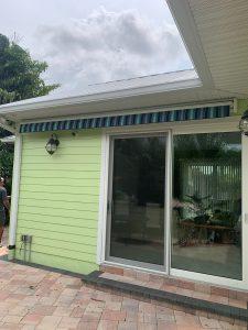 A house with green siding features a sliding glass door shaded by a striped blue and green awning. The patio is paved with light-colored bricks, and theres a leafy tree nearby. Two outdoor light fixtures flank the door, and the sky is overcast.