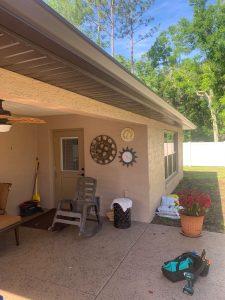 A patio with a rocking chair, broom, and various clocks on the wall. A potted plant with red flowers is on the right. A set of tools is on the ground near a wicker stool. The area is shaded under a roof, with trees in the background.