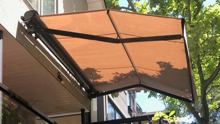 A brown retractable awning extends over a balcony, casting shade. Tree branches and leaves create dappled shadows on the fabric. Nearby buildings are partially visible in the background.