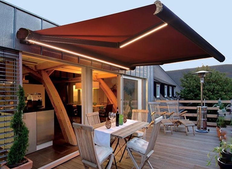 A modern outdoor patio with a retractable, brown awning featuring built-in lights. Wooden furniture surrounds a dining table set for a meal, and the area overlooks other buildings.